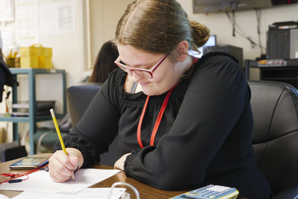 A female student seated at a desk completing a math assignment with an Orion TI calculator and Iphone both visible on the table. 