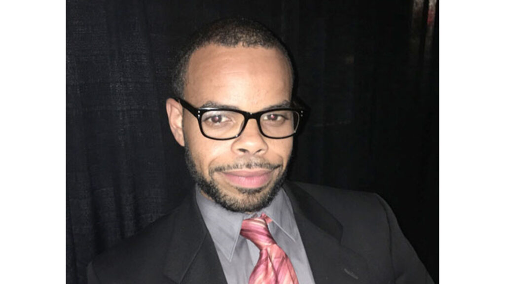 An Afro-Latino man with a short haircut and beard wearing black-framed glasses smiles for the camera. He’s wearing a black suit jacket with a shirt and tie.