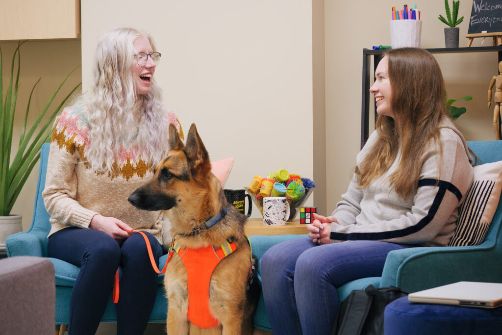  Two individuals sit and talk in a colorful office. One individual holds onto the leash of a dog guide. 