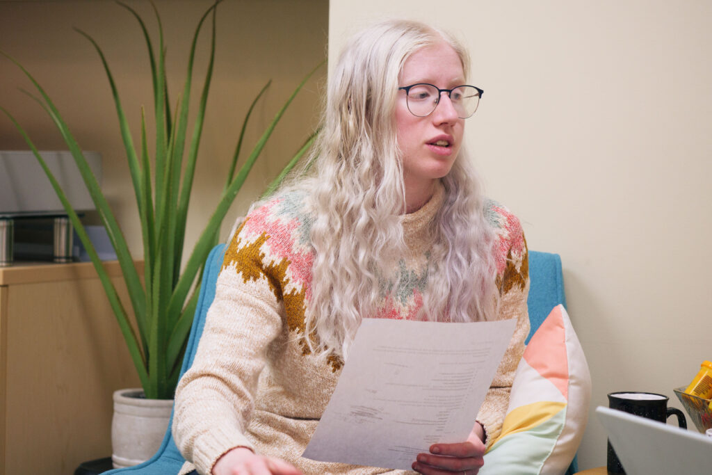 Person with albinism (fair skin, blonde hair, and glasses) sits on a chair and holds a paper.